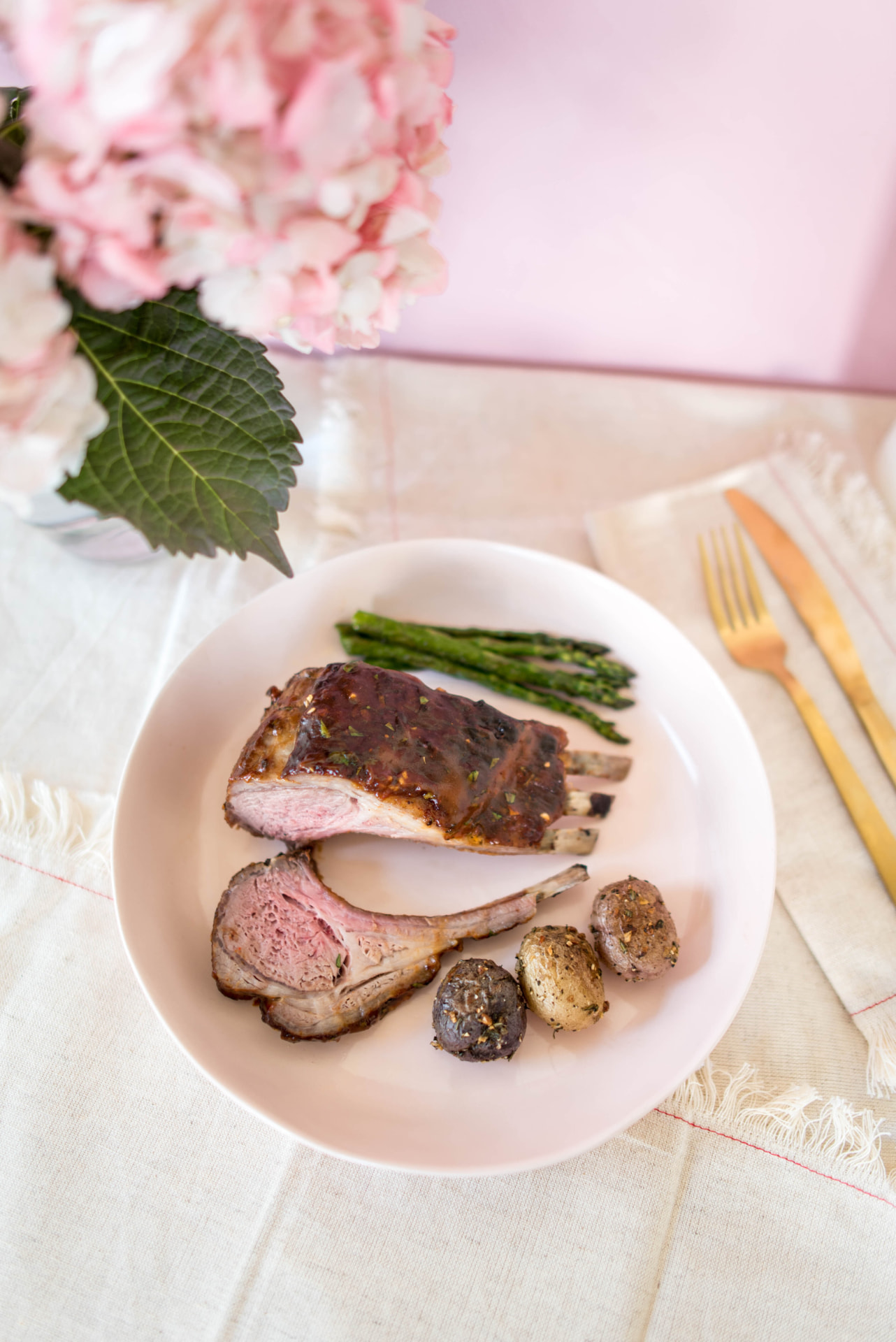 A plate with roasted lamb, asparagus, and potatoes beside pink hydrangeas on a spring table